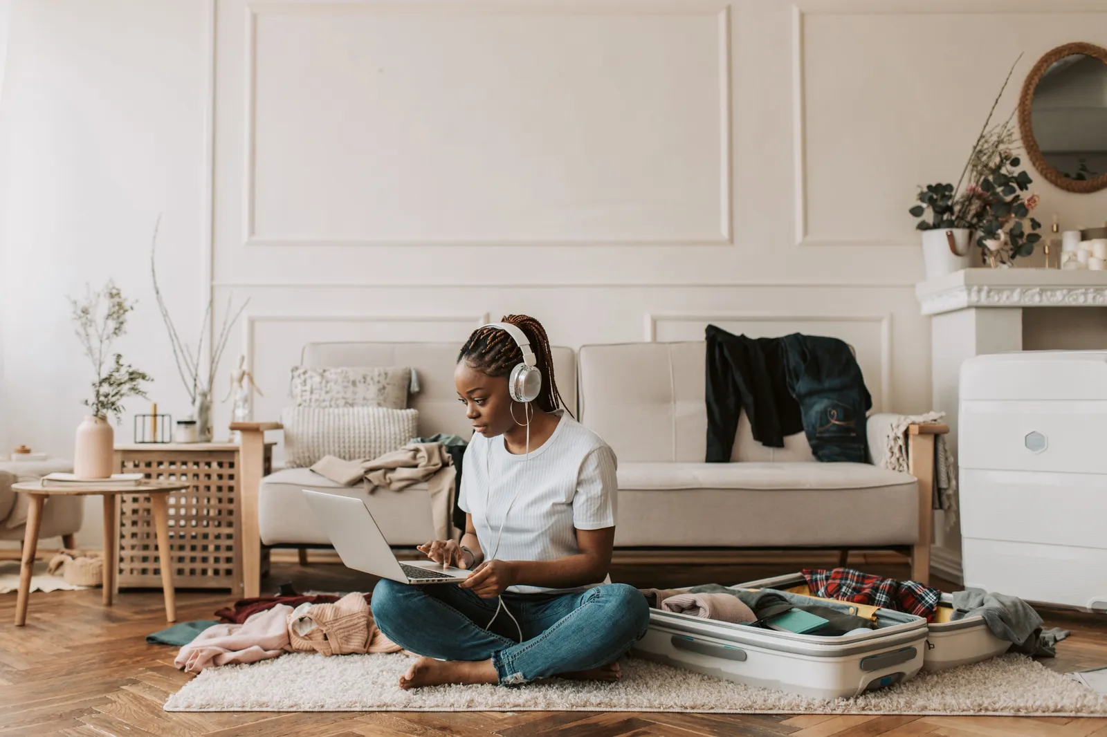 femme assise en tailleur dans son salon, ordinateur portable sur les genoux. elle travaille avec une valise ouverte et remplie à moitié à côté d'elle
