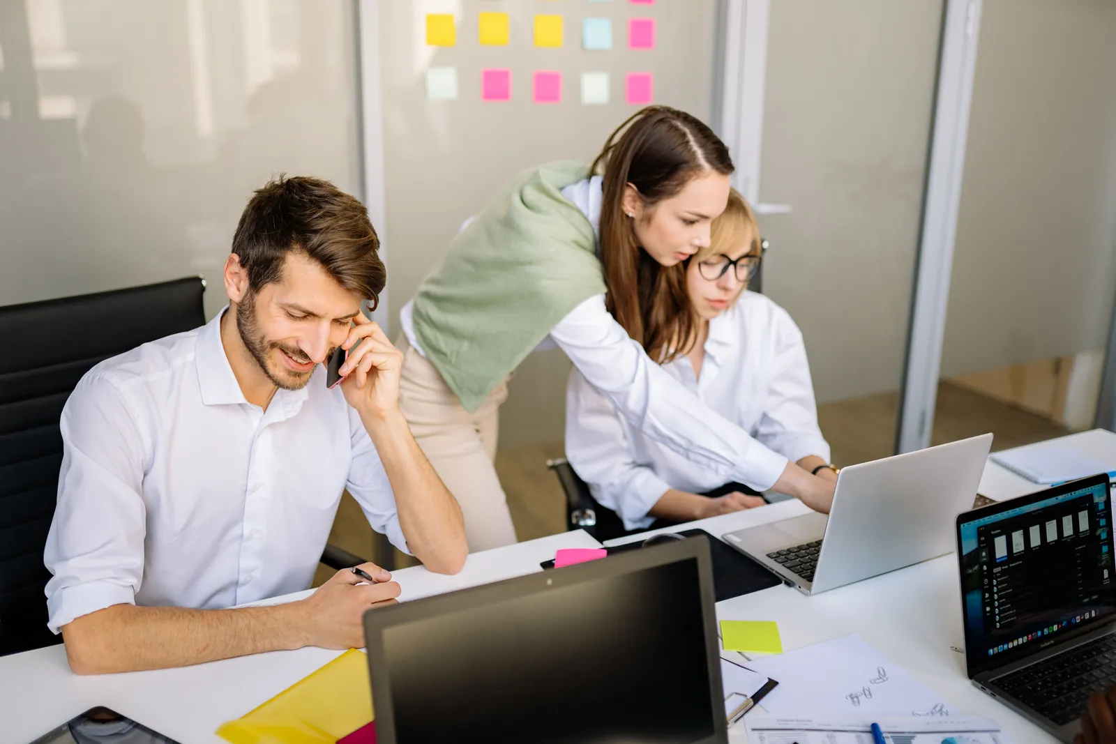 une équipe formée de 2 femmes et un homme travaillent dans un bureau. L'homme est au téléphone et l'une des femmes montre à l'autre quelque chose sur l'écran de l'ordinateur. Sur le mur vitré dans le dos des personnes il y a des post-its colorés
